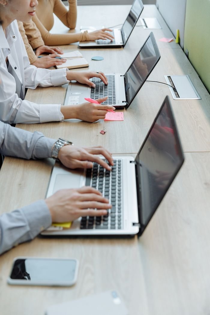 About Group of office workers typing on laptops at a shared workspace desk.