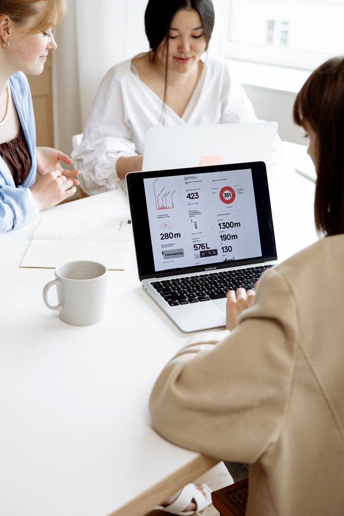 Three women discussing business strategies at a desk while showing data on a laptop in a bright office.